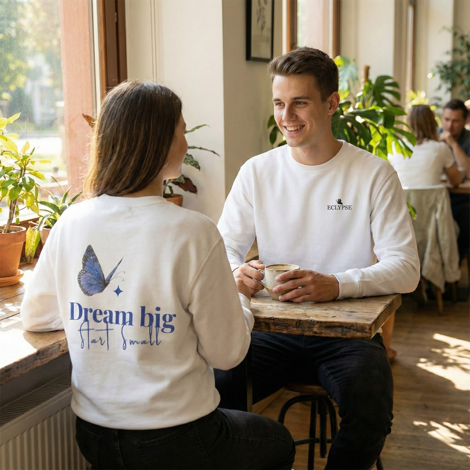 Een lachende man en vrouw drinken koffie in witte Eclypse sweaters met vlinderprint en de tekst Dream big start small.